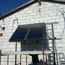 Montaż paneli solarnych na ścianie budynku z białej cegły, widoczne rusztowanie i drabina, instalacja w trakcie realizacji.