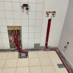 Wnętrze łazienki w trakcie prac instalacyjnych: czerwone rury rozprowadzone po podłodze i ścianach, widoczne otwory w ścianach z izolacją piankową i instalacjami elektrycznymi, częściowo ułożone...