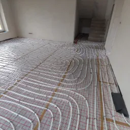 Białe rurki ogrzewania podłogowego ułożone na czerwonej siatce w surowym wnętrzu z oknem i schodami w tle.