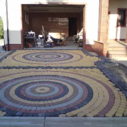 Kolorowa nawierzchnia z kostki brukowej ułożona w dwa koncentryczne okręgi przed garażem i wejściem do domu.