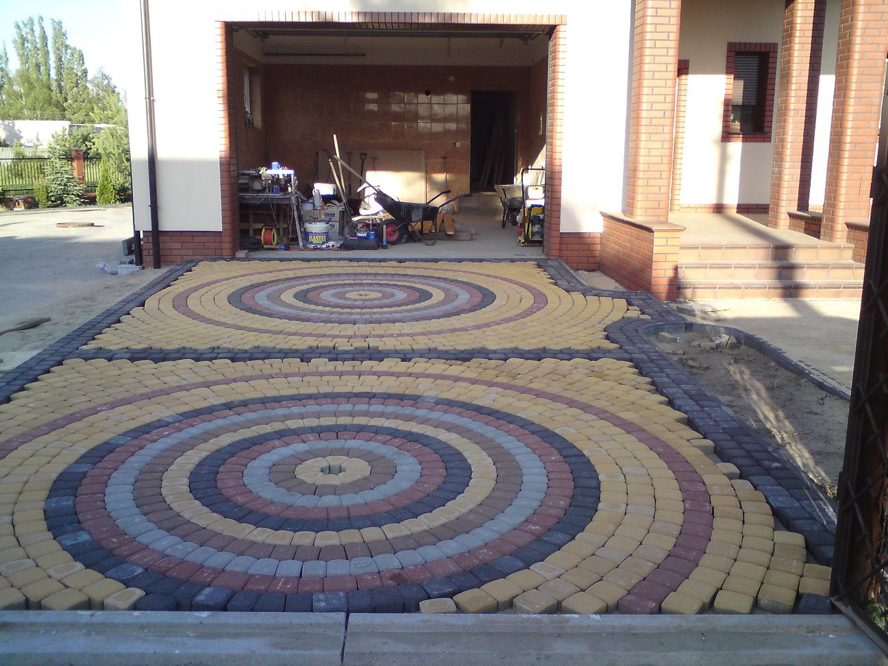Kolorowa nawierzchnia z kostki brukowej ułożona w dwa koncentryczne okręgi przed garażem i wejściem do domu.