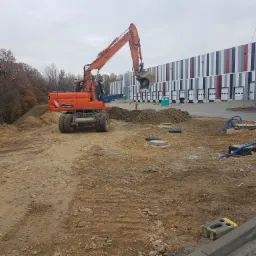 Pomarańczowa koparka kołowa na placu budowy, z tyłu hala magazynowa z bramami, ziemia rozkopana i nierówna, słaba widoczność.