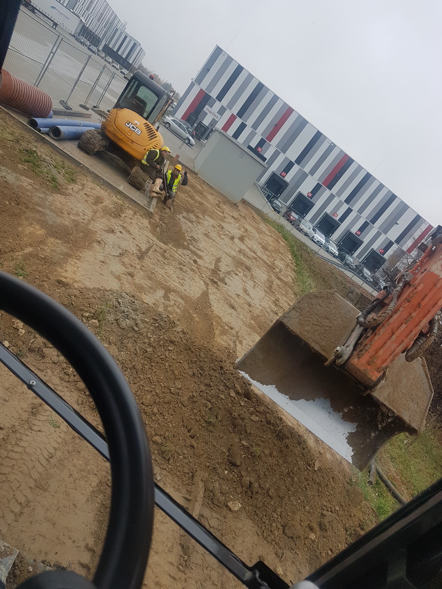 Widok z kabiny koparki na wykop, w tle minikoparka JCB i pracownicy w kamizelkach odblaskowych, obok składowane rury, za ogrodzeniem hala magazynowa.