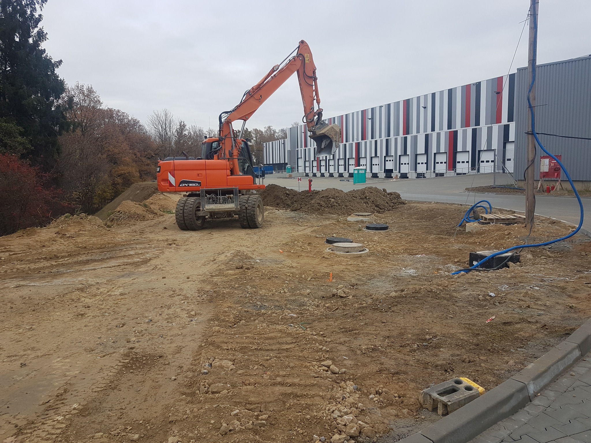 Pomarańczowa koparka kołowa na placu budowy, z tyłu hala magazynowa z bramami, ziemia rozkopana i nierówna, słaba widoczność.