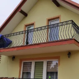 Żółta elewacja domu z balkonem z ozdobną, czarną, metalową balustradą o falistym wzorze. Na balkonie widoczna niebieska plandeka. Pod balkonem okna z roletami w kolorze brązowym.