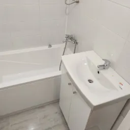 Biała łazienka po remoncie: wanna, umywalka z szafką, chromowana bateria i białe płytki na ścianach.