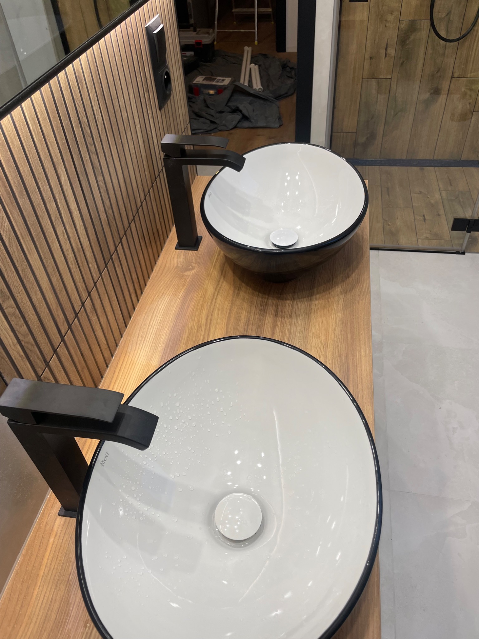 Dwie nowoczesne umywalki nablatowe z czarnymi bateriami na drewnianym blacie. W tle widoczna ściana z drewnianymi panelami i fragment kabiny prysznicowej.