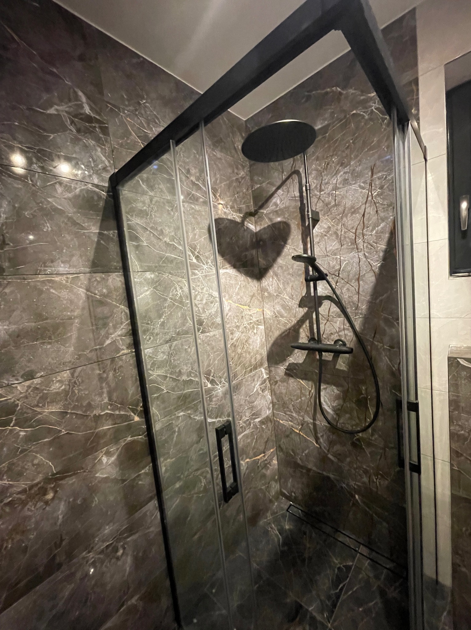 Nowoczesna kabina prysznicowa z czarnymi profilami i deszczownicą, wykończona płytkami imitującymi marmur. Minimalistyczny design, eleganckie detale.