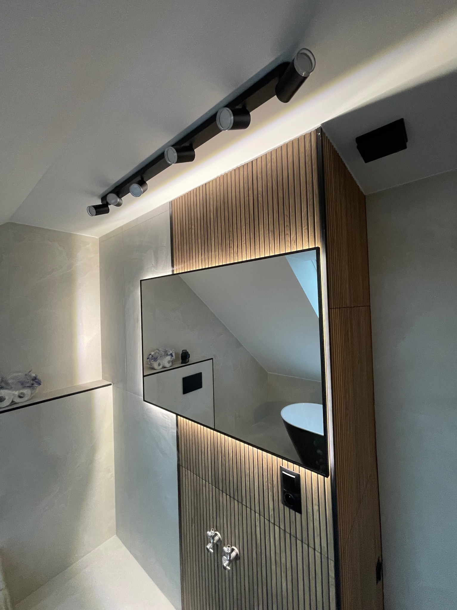 Nowoczesna łazienka z podświetlanym lustrem, drewnianymi panelami i punktowym oświetleniem sufitowym. W lustrze odbija się fragment wanny.