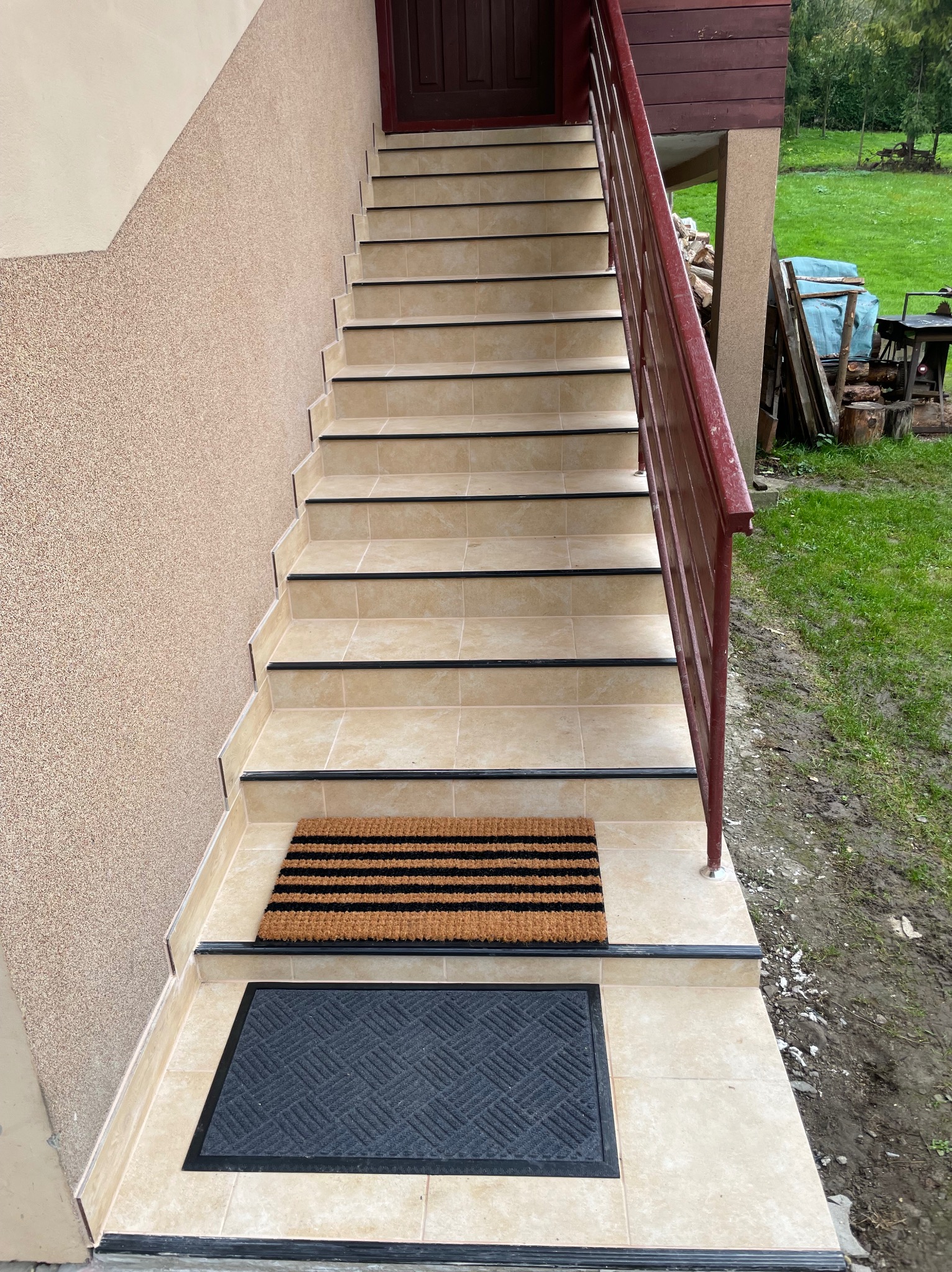 Wyłożone beżowymi płytkami schody zewnętrzne z brązową balustradą, prowadzące do ciemnych drzwi. Na dole dwie wycieraczki: brązowa i czarna. Tynk strukturalny na ścianie.