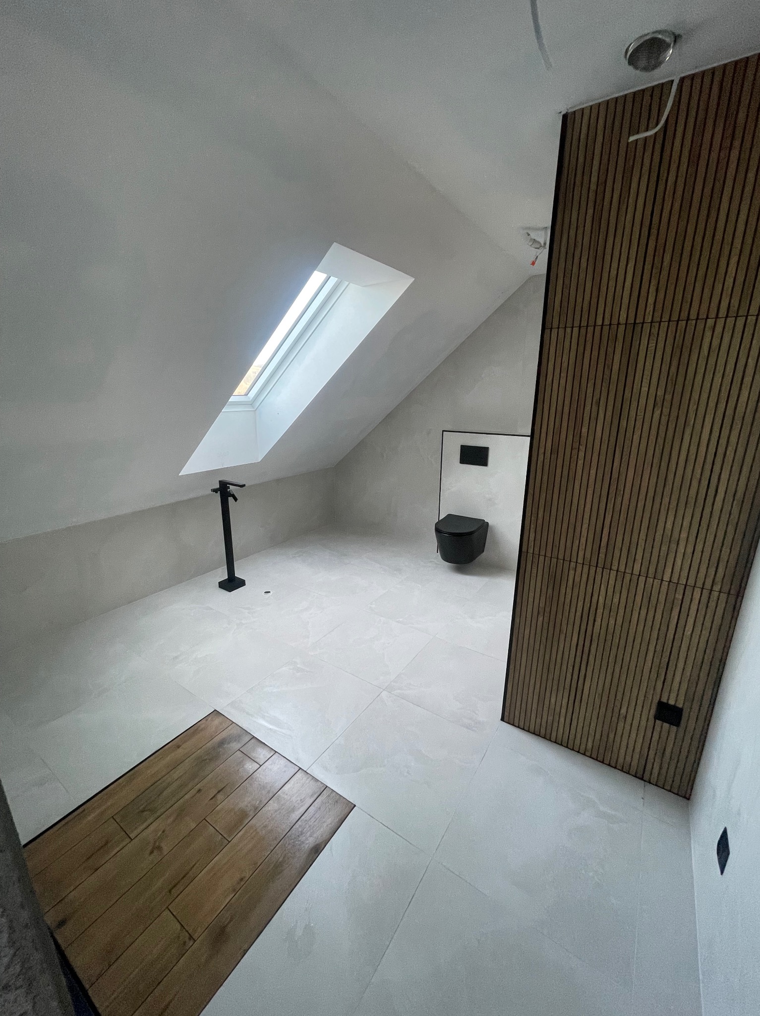Nowoczesna łazienka na poddaszu z oknem dachowym, czarną armaturą i drewnianą ścianą. Minimalistyczny design, jasne płytki podłogowe, skosy.