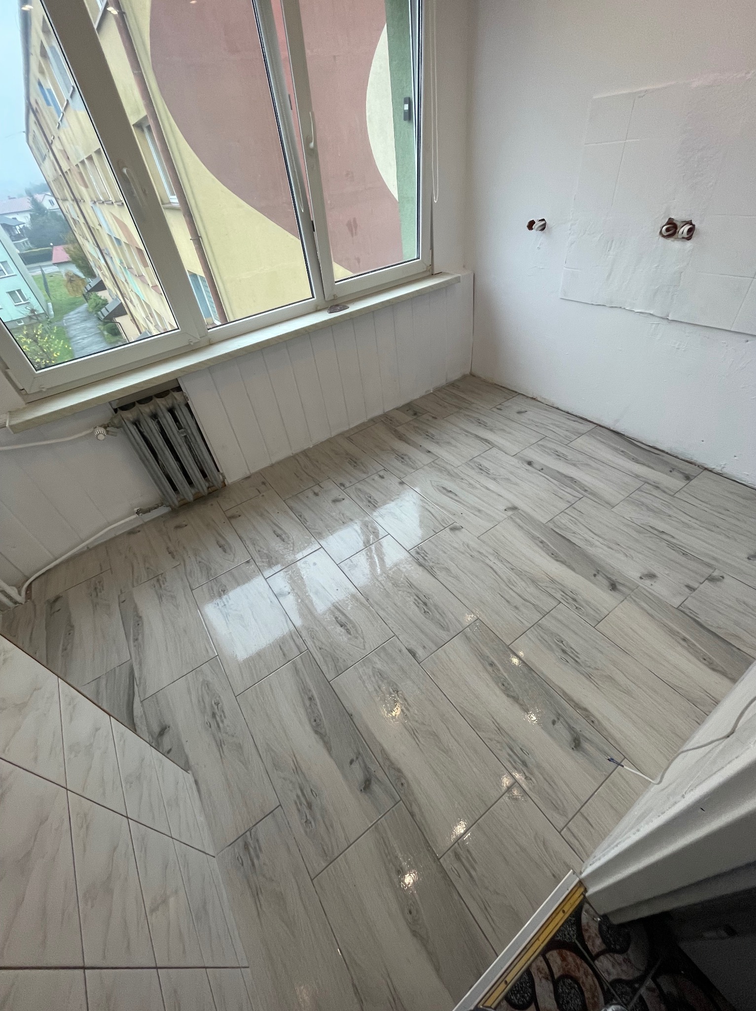 Jasne, drewnopodobne płytki podłogowe ułożone w nowo wyremontowanym pokoju z oknem i grzejnikiem. Widoczne puszki elektryczne na ścianie.