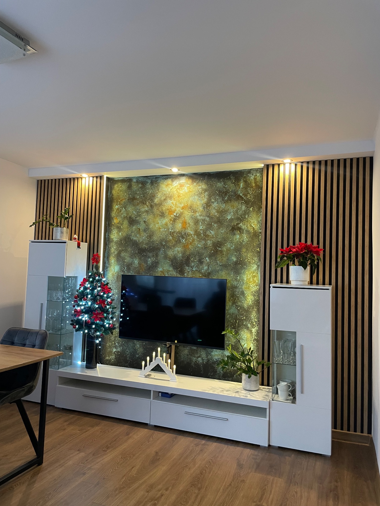 Salon z telewizorem na tle ściany z drewnianych lameli i dekoracyjnej tapety. Oświetlenie punktowe, białe meble, świąteczne dekoracje i drewniana podłoga.