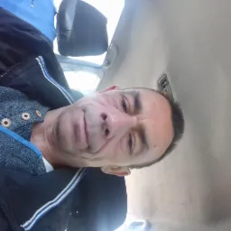 Mężczyzna w samochodzie, ubrany w sweter i kurtkę, spogląda w obiektyw. Widoczna podsufitka auta i zagłówek.