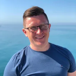 Uśmiechnięty mężczyzna w okularach i niebieskiej koszulce, z morzem w tle, prezentuje się profesjonalnie i budzi zaufanie.