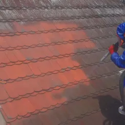 Pracownik w kombinezonie ochronnym czyści dachówkę za pomocą specjalistycznego sprzętu, widoczny efekt czyszczenia na części dachu.