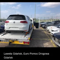 Biały samochód Volkswagen Golf GTI na żółtej lawecie, otwarte drzwi lawety, parking z innymi samochodami w tle, błękitne niebo z chmurami.