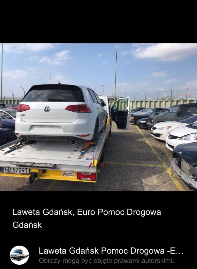 Biały samochód Volkswagen Golf GTI na żółtej lawecie, otwarte drzwi lawety, parking z innymi samochodami w tle, błękitne niebo z chmurami.