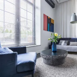 Nowoczesny salon z dużym oknem przesłoniętym poziomymi listwami, niebieski fotel i sofa, abstrakcyjny obraz na ścianie.