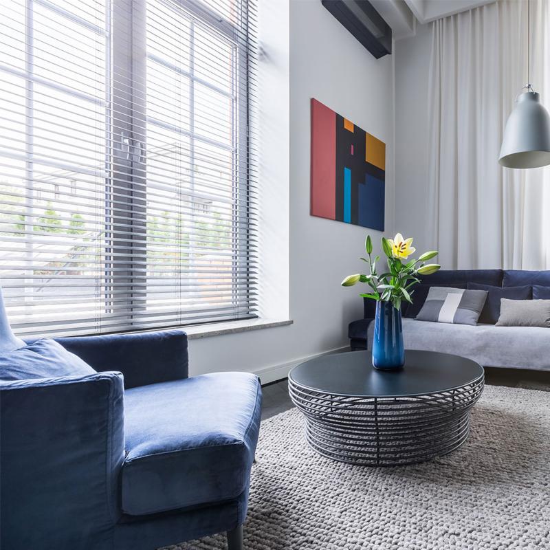 Nowoczesny salon z dużym oknem przesłoniętym poziomymi listwami, niebieski fotel i sofa, abstrakcyjny obraz na ścianie.