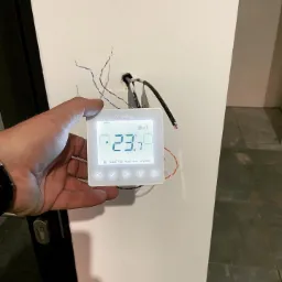 Ręka trzyma biały, dotykowy panel sterowania Heatmiser, zamontowany na ścianie, z widocznymi przewodami instalacyjnymi; wyświetlacz pokazuje temperaturę 23.7 stopni Celsjusza.