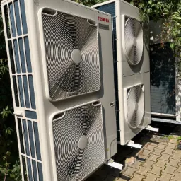 Dwa zewnętrzne moduły klimatyzacji Toshiba Digital Inverter zamontowane na białych wspornikach, na tle zieleni i kostki brukowej.