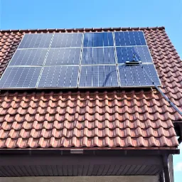 Czyszczenie paneli słonecznych na dachu domu z czerwoną dachówką za pomocą szczotki na długim wysięgniku, błękitne niebo w tle.