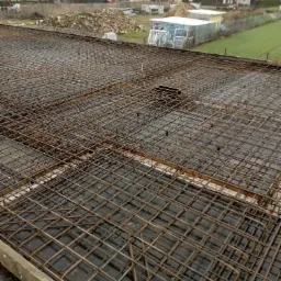 Zbrojenie płyty fundamentowej domu jednorodzinnego, widoczne stalowe pręty ułożone w kratę na czarnej folii izolacyjnej, w tle zabudowa mieszkalna.