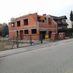 Surowy dwupiętrowy budynek w trakcie budowy, z czerwonej cegły i betonowych nadproży, widoczny z ulicy za ogrodzeniem, obok zaparkowany różowy samochód.