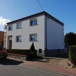 Dwukondygnacyjny budynek z jasną elewacją i szarymi akcentami, widoczny z poziomu ulicy, z zaparkowanym pomarańczowym vanem i fragmentem rusztowania po lewej stronie.