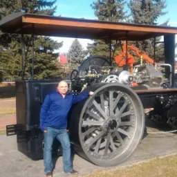 Mężczyzna opiera się o zabytkowy walec parowy z odkrytym silnikiem, eksponowany na zewnątrz na betonowym postumencie, otoczony drzewami i budynkami w tle.