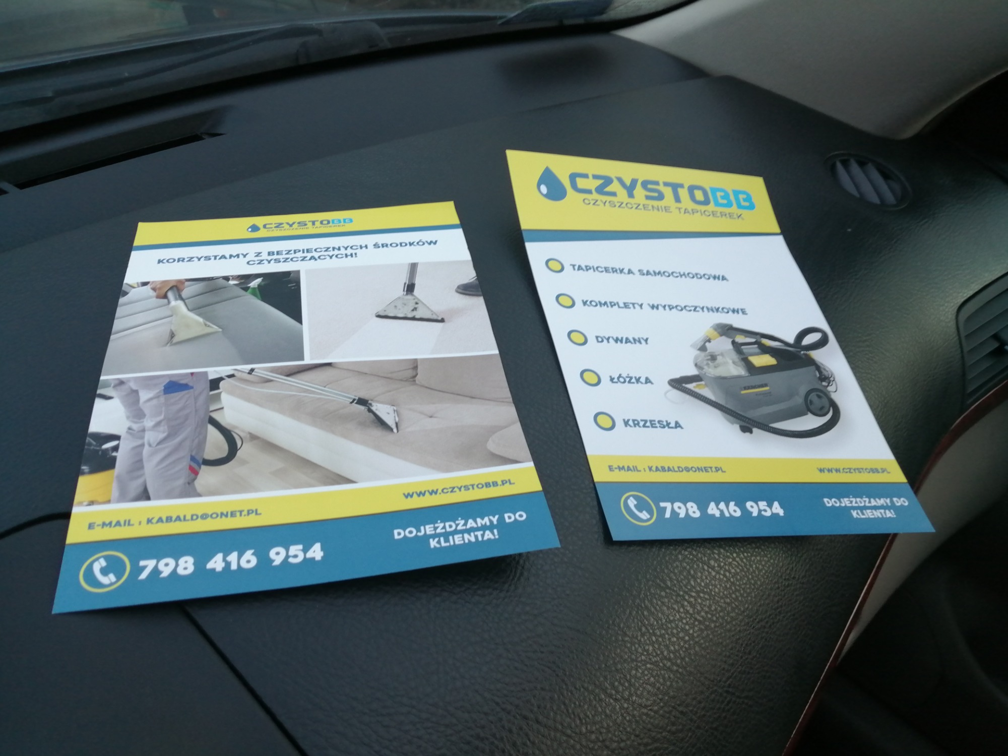 Dwie ulotki reklamowe firmy oferującej usługi czyszczenia tapicerki, leżące na desce rozdzielczej samochodu. Na ulotkach widoczne zdjęcia i informacje kontaktowe.
