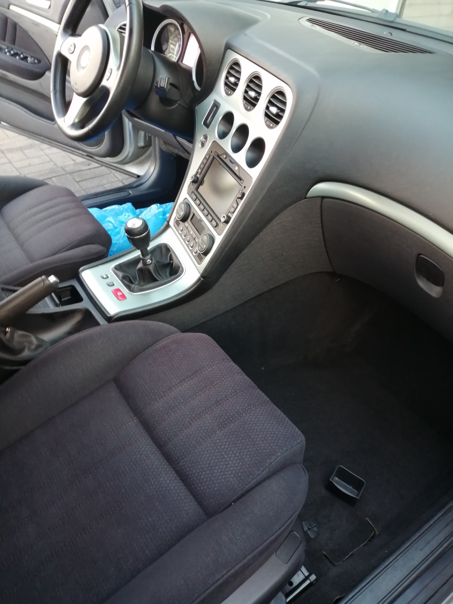 Wnętrze samochodu Alfa Romeo z widocznymi czarnymi, tekstylnymi fotelami, deską rozdzielczą w kolorze czarnym i srebrnym oraz elementami sterowania. Na podłodze widoczne drobne śmieci i plastikowy...