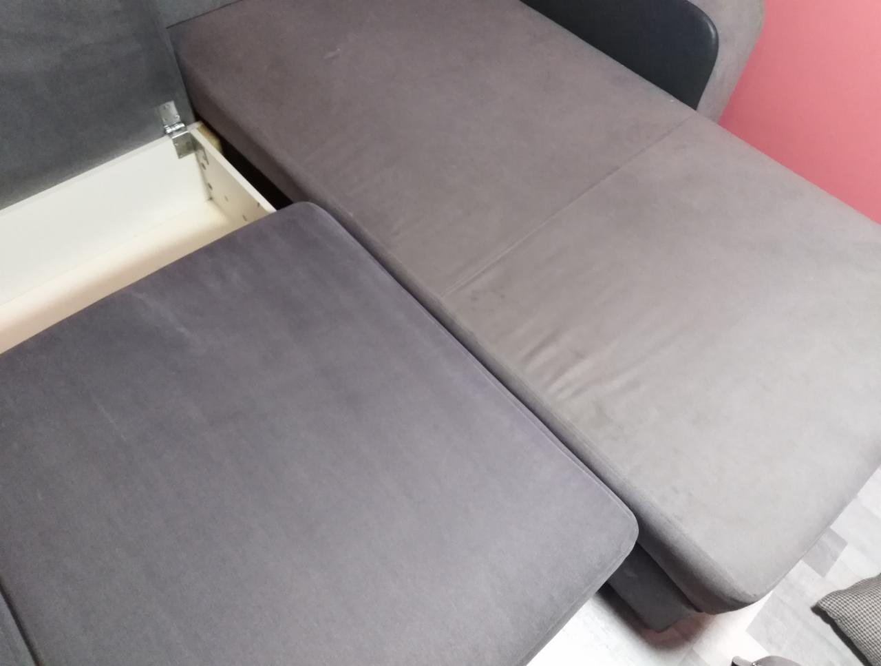 Szara, rozłożona kanapa z wysuwanym pojemnikiem na pościel, widoczne zawiasy i wewnętrzna, biała część pojemnika.