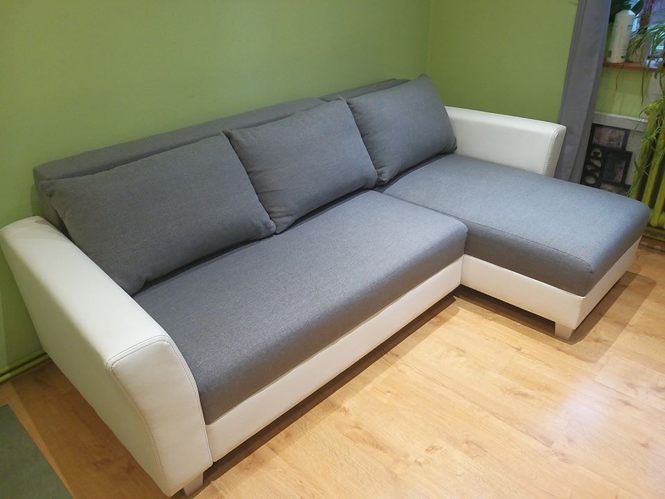 Szaro-biała narożna kanapa z trzema poduszkami na drewnianej podłodze, widok z niskiego kąta, z zieloną ścianą w tle.