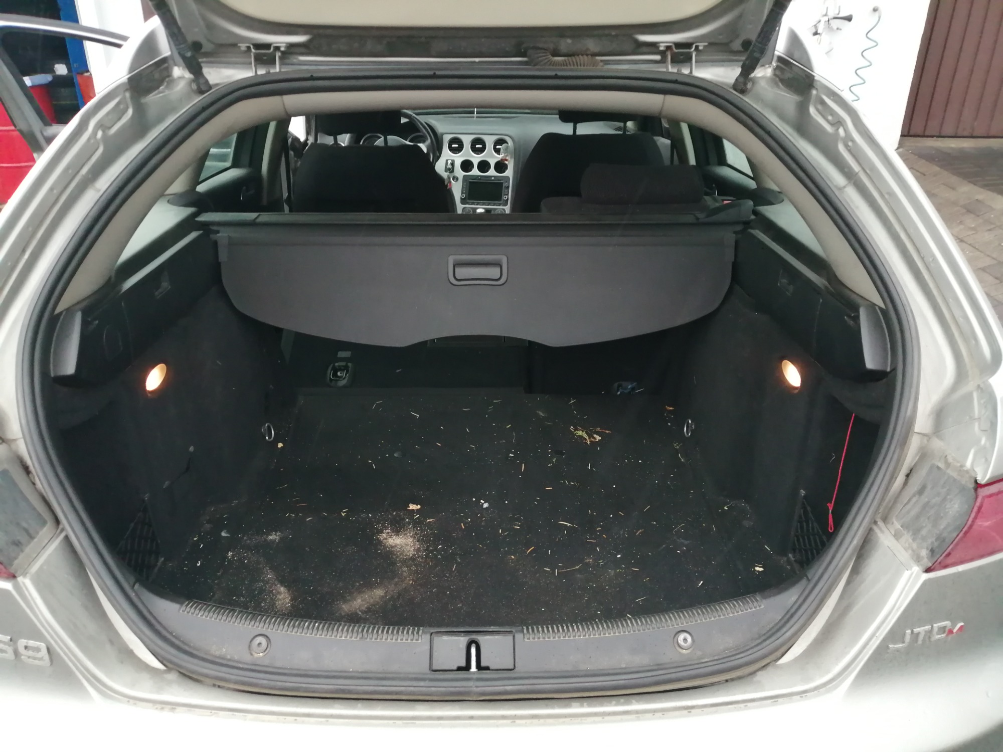 Bagażnik srebrnego kombi Alfa Romeo 159 z zasłoniętą roletą, widoczne drobne zabrudzenia na wykładzinie.