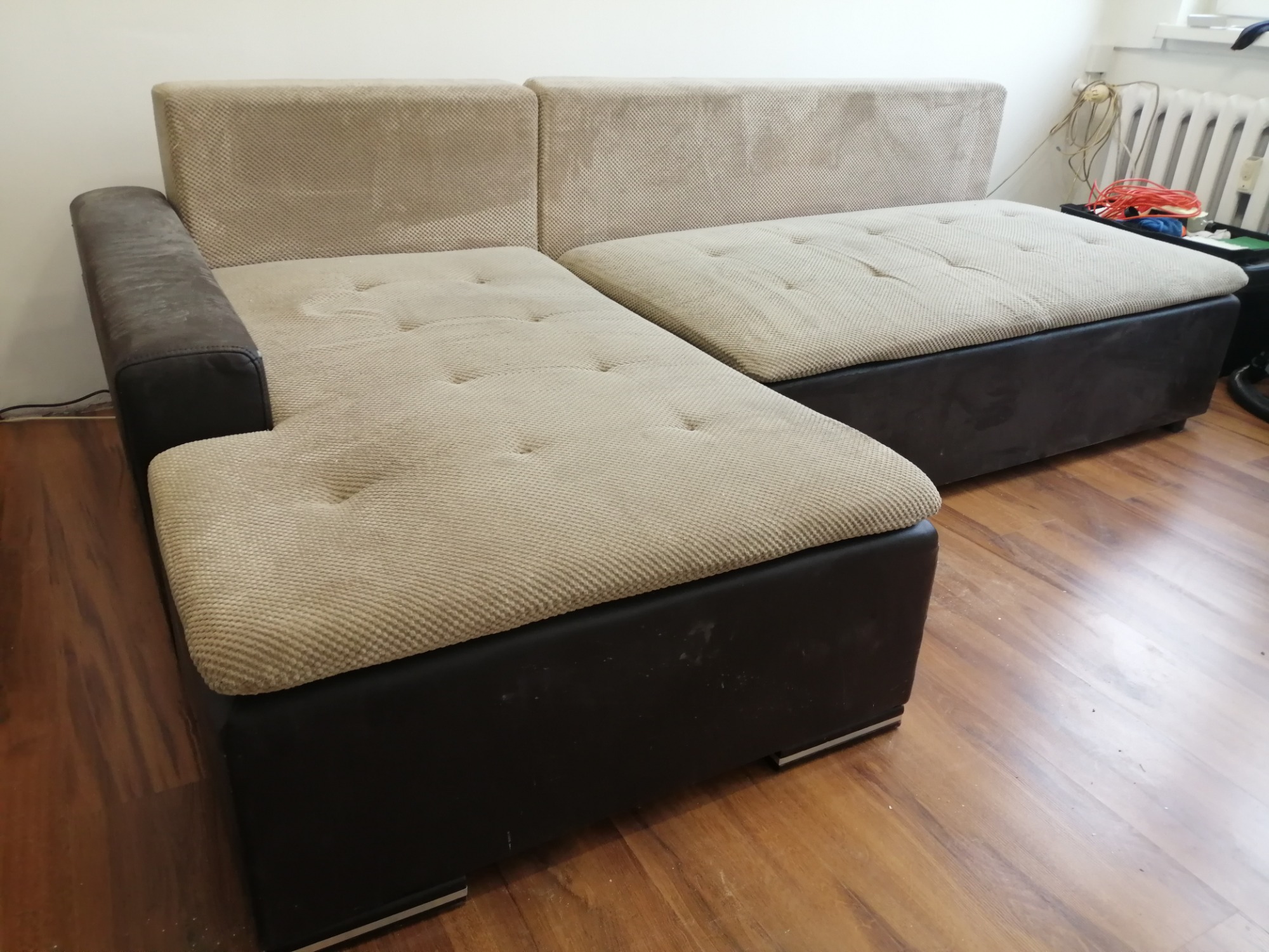 Narożnik z beżową tapicerką w odcieniach brązu i ciemnobrązową podstawą, widoczne plamy na boku, drewniana podłoga