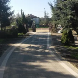 Ukończony podjazd z szarej kostki brukowej z białymi obrzeżami prowadzący do bramy wjazdowej, w tle widoczny dom i drzewa.