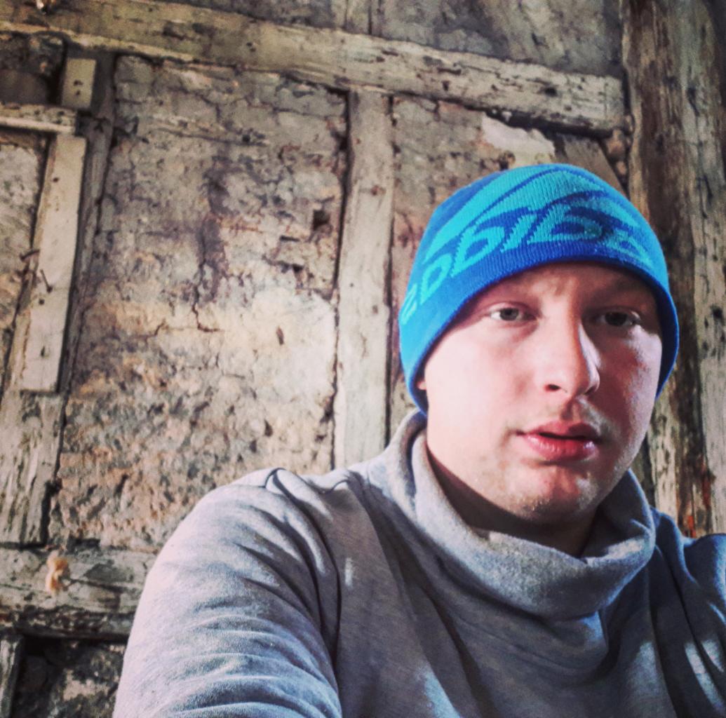 Mężczyzna w niebieskiej czapce na tle ściany z surowego drewna i szarego tynku o nierównej fakturze.