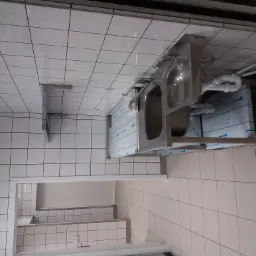 Profesjonalna, dwukomorowa umywalka ze stali nierdzewnej zamontowana w pomieszczeniu wyłożonym białymi płytkami, widoczne rury odpływowe i elementy mocujące, zabezpieczone folią ochronną.