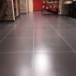 Wypolerowana, ciemnoszara podłoga z płytek w lokalu komercyjnym, widoczne fugi, fragment ceglanej ściany i elementy wyposażenia wnętrza.