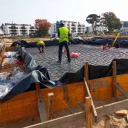 Przygotowanie zbrojenia i szalunku pod fundament płyty budynku mieszkalnego, widoczne elementy konstrukcyjne i pracownicy w odblaskowych kamizelkach.