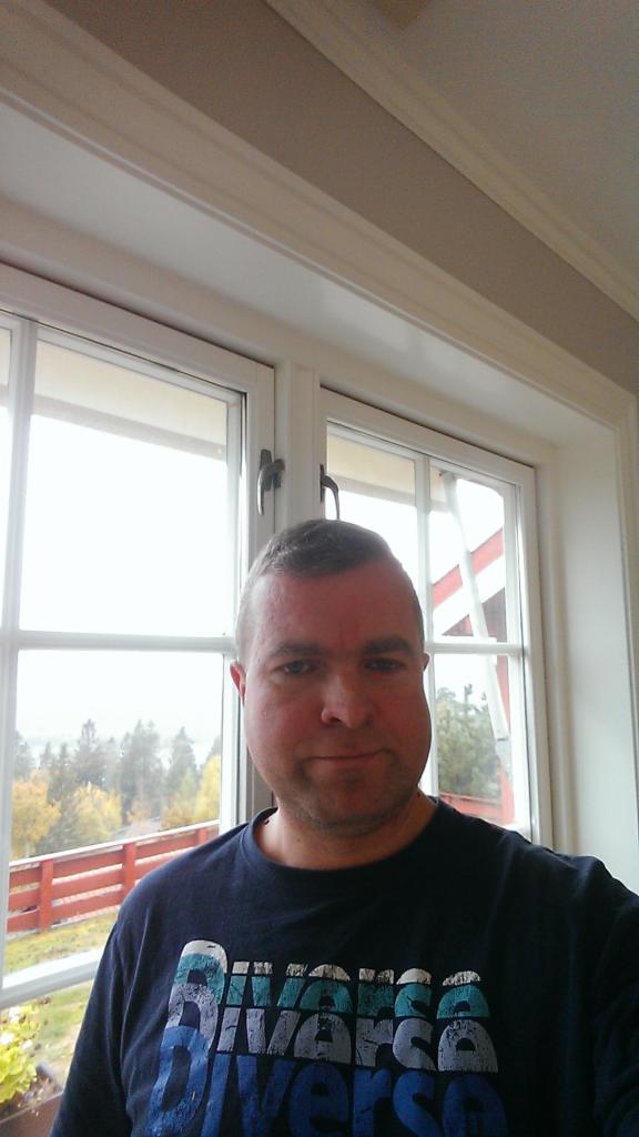 Mężczyzna w niebieskiej koszulce z nadrukiem stoi na tle białego okna z widokiem na zewnątrz. Za oknem widoczne drzewa i czerwone ogrodzenie.