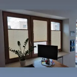 Trzy okna z drewnianymi ramami, osłonięte kremowymi plisami, częściowo zasłonięte, z widokiem na budynki na zewnątrz; na parapecie roślina doniczkowa i monitor komputerowy.