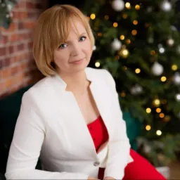 Portret uśmiechniętej kobiety w białej marynarce i czerwonej sukience, siedzącej na zielonej sofie na tle ceglanej ściany i choinki z bombkami.