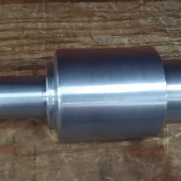 Metalowy, symetryczny element obrotowy, prawdopodobnie wał lub trzpień, po obróbce, leżący na drewnianym blacie.