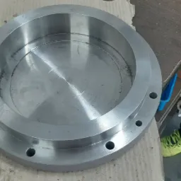 Obrabiany metalowy element w kształcie pierścienia z otworami, leżący na kartonie, widoczne ślady obróbki.