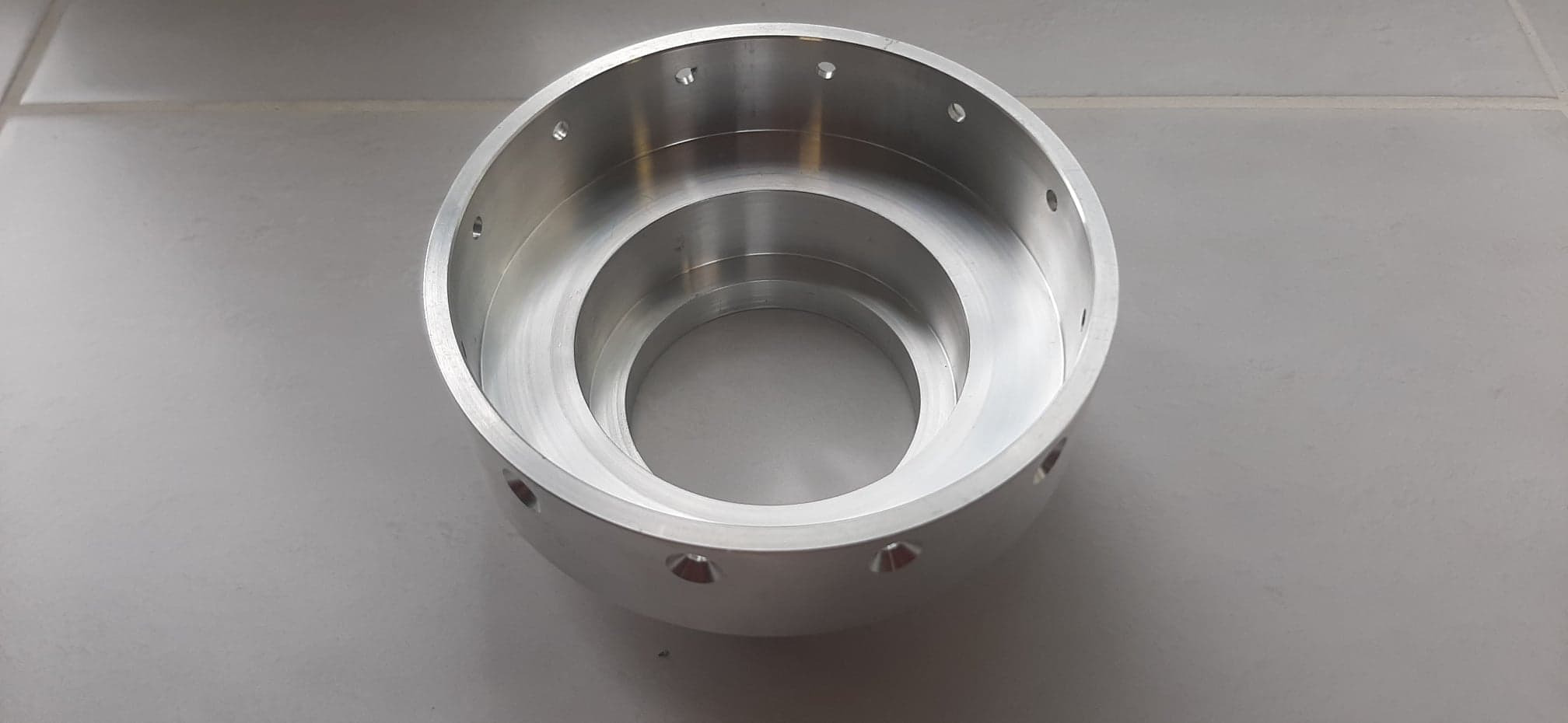 Aluminiowy element toczony z otworami, leżący na szarej płytce, widok z góry.