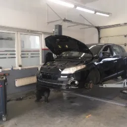 Czarny samochód na podnośniku w warsztacie, z otwartą maską i zdjętym kołem, widoczny mechanik pracujący pod autem.