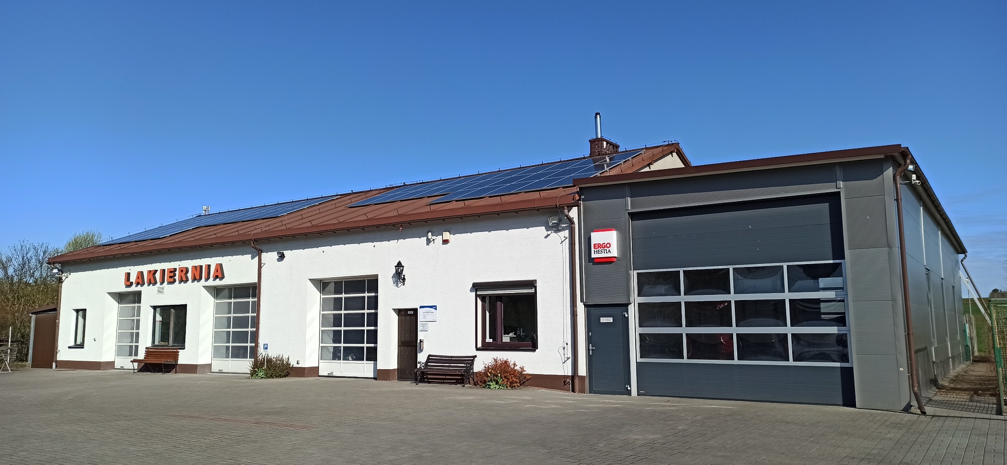Budynek warsztatu z napisem 'LAKIERNIA' i panelami słonecznymi na dachu, obok nowoczesna dobudówka z szarą bramą segmentową.